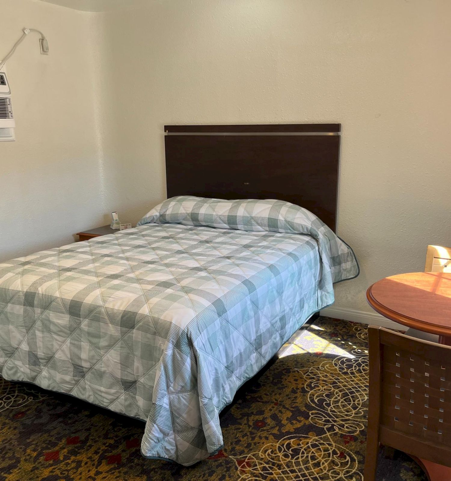 A simple room with a bed, an air conditioner, a small table, chairs, a lamp, and a patterned carpet.