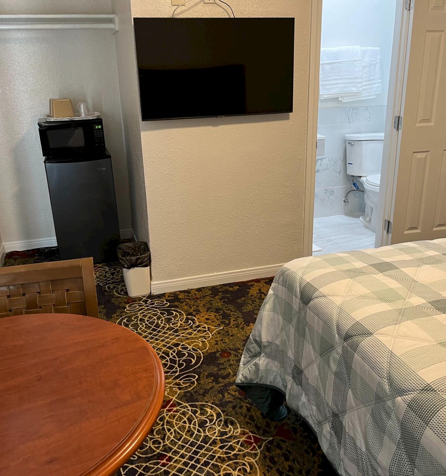 A hotel room with a bed, round table, chair, wall-mounted TV, closet, mini-fridge, and a bathroom in the background.