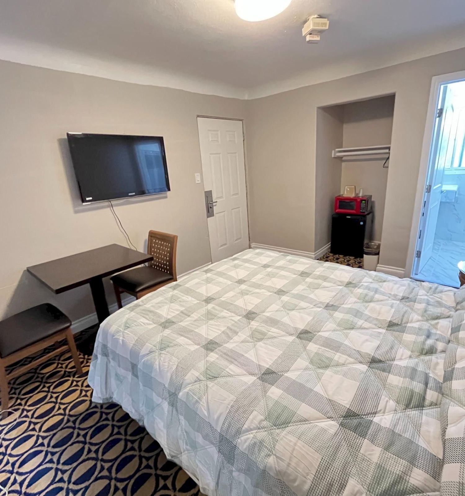 The image shows a small hotel room with a bed, TV, table, chairs, open closet, and adjacent bathroom.