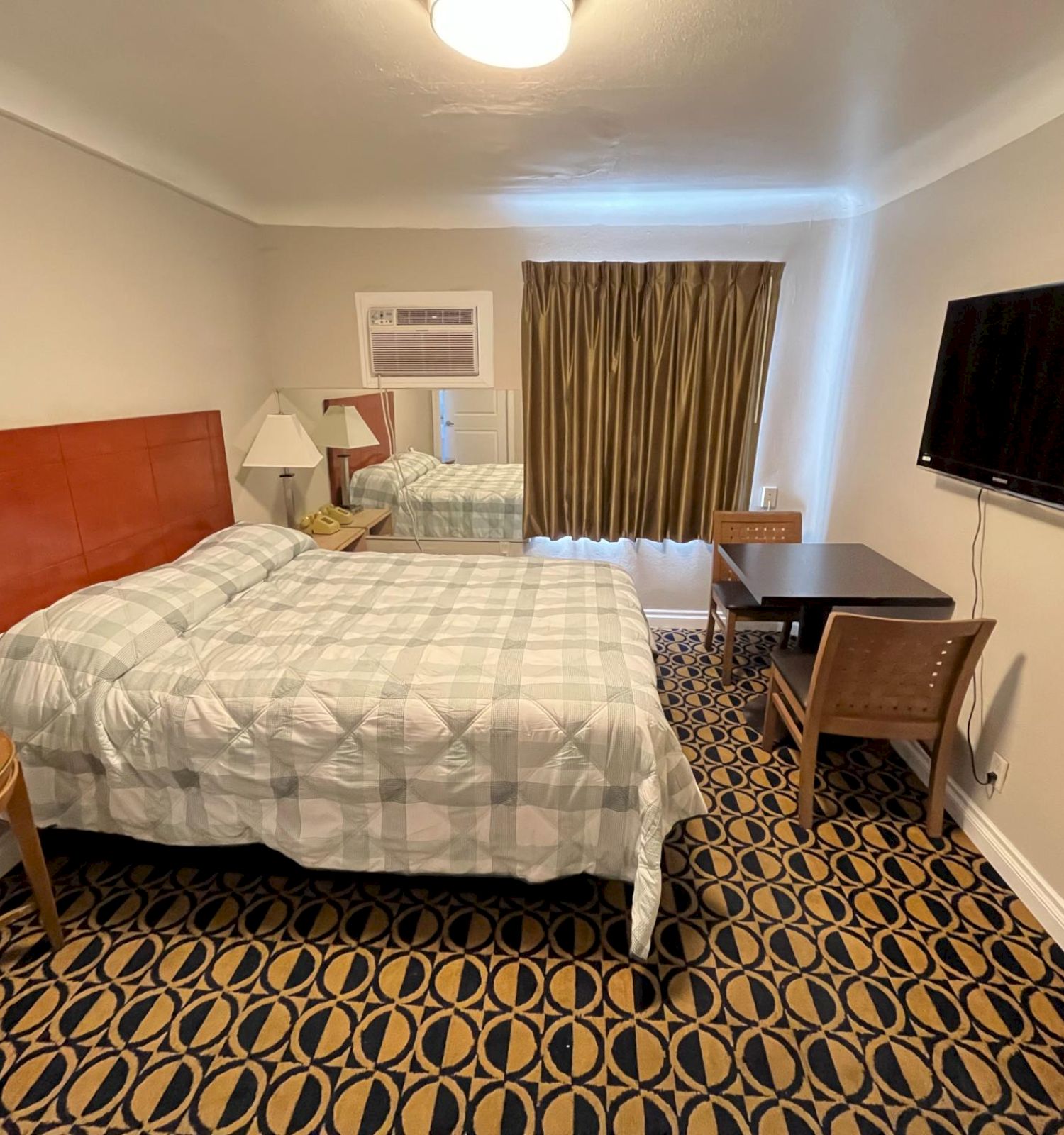 A hotel room with a bed, nightstands, lamps, a mounted TV, table, chairs, and patterned carpet. Simple and functional interior design.