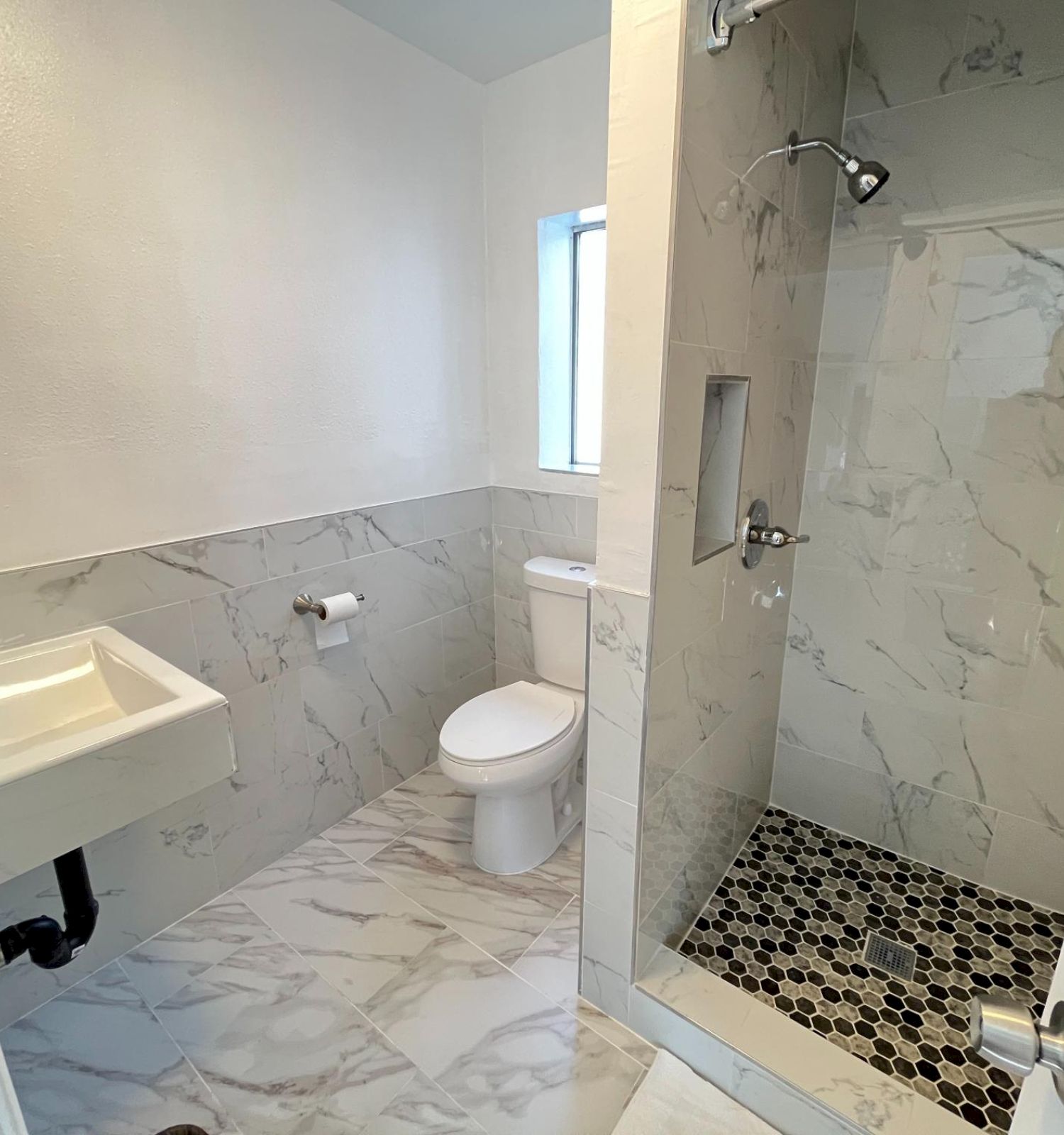 The image shows a bathroom with a sink, toilet, and a shower with marbled tiles and a pebble floor design.