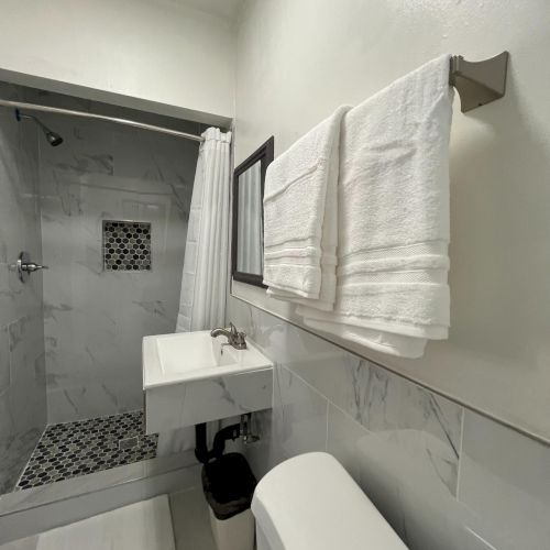 A modern bathroom with a shower, toilet, sink, and white towels on a rack. The design is clean and minimalistic.