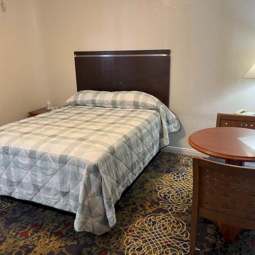 A hotel room with a bed, checked bedding, a round table with two chairs, a lamp, and an air conditioning unit on the wall.