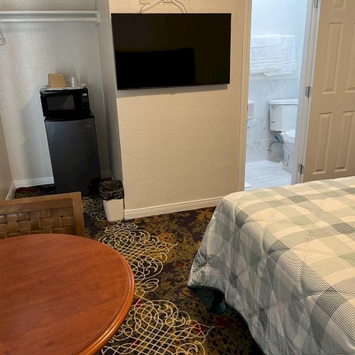 The image shows a hotel room with a bed, wall-mounted TV, mini fridge, and a bathroom. A small round table is also in view.