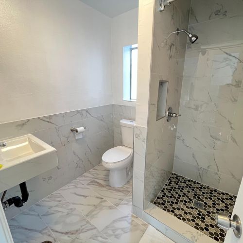 The image shows a modern bathroom with a sink, toilet, and a shower featuring a marble design and pebble-tiled floor.