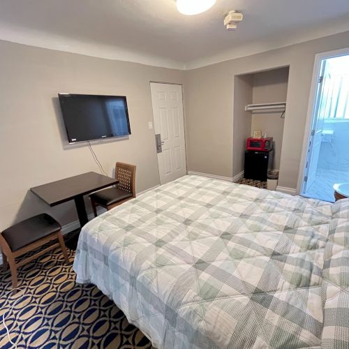 The image shows a hotel room with a bed, wall-mounted TV, table, chairs, and an open bathroom door. The decor is simple and neat.