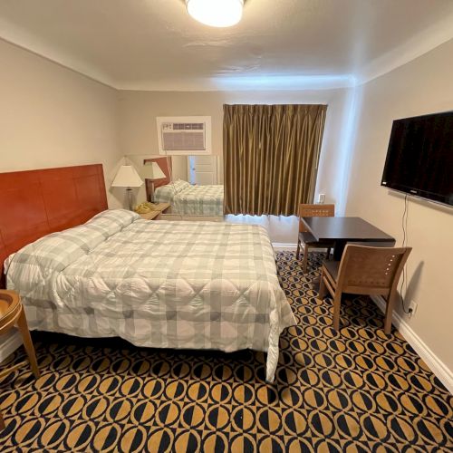 The image shows a hotel room with a bed, TV, desk, chairs, and a window with curtains. The floor has patterned carpeting.