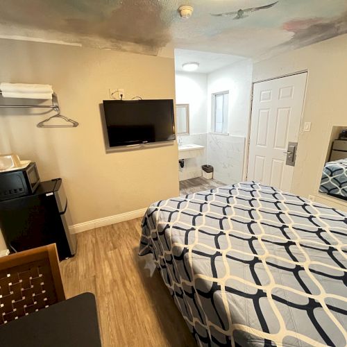 This image shows a small hotel room with a bed, TV, mini fridge, and bathroom. The decor is simple with wooden flooring.