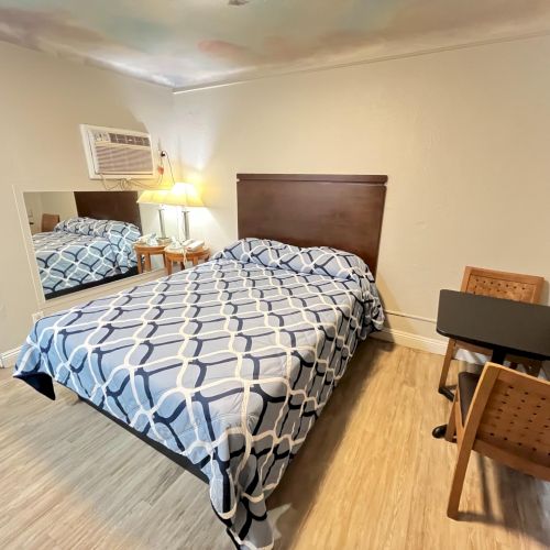 A basic hotel room with a bed, patterned bedspread, small table with two chairs, a lamp, wall mirror, and window air conditioner unit.