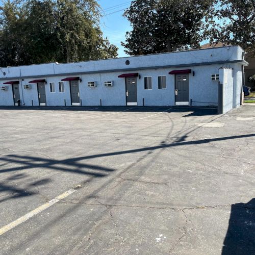 The image shows a single-story building with several doors, possibly a motel, and an empty parking area in front of it.