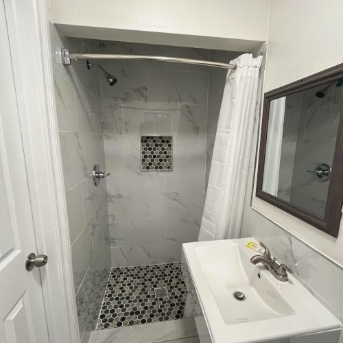 The image shows a small bathroom with a walk-in shower, white tiles, a corner sink, and a mirror above it.