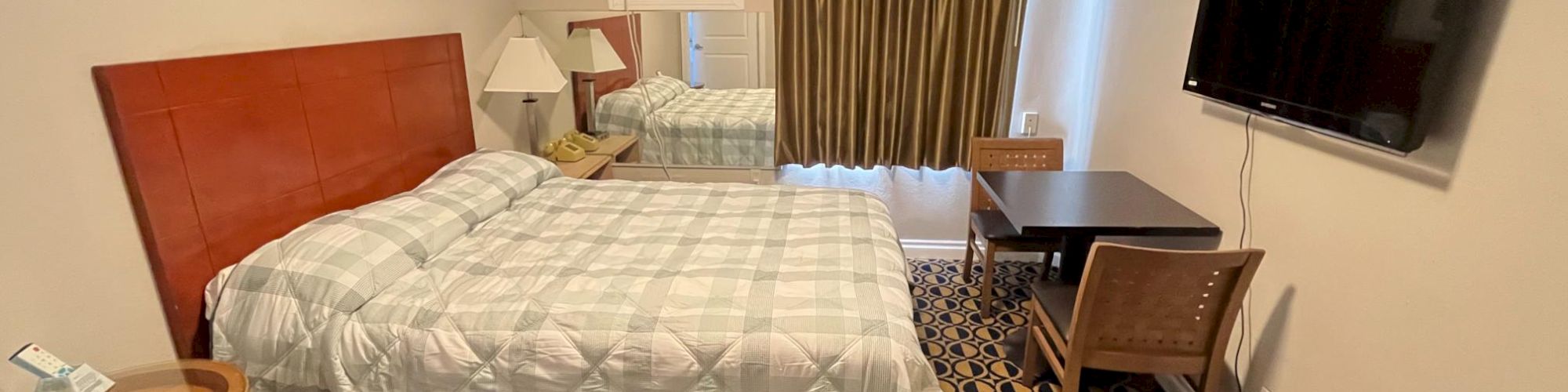 A hotel room with a neatly made bed, a TV on the wall, a desk and chair, patterned carpet, and a window with curtains.