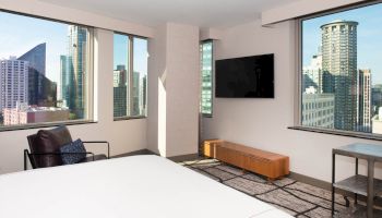 The image shows a modern room with large windows offering a cityscape view, a wall-mounted TV, a bed, and a chair.
