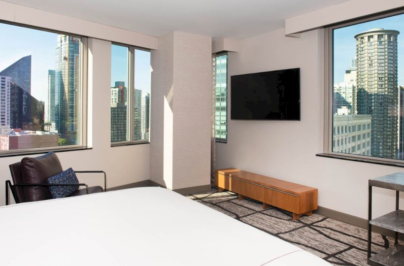 The image shows a modern room with large windows offering a cityscape view, a wall-mounted TV, a bed, and a chair.
