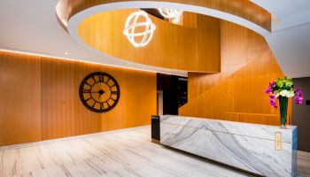 A modern, stylish lobby with a marble reception desk, wooden accents, a large wall clock, spiral design element, and flower vase on the desk.