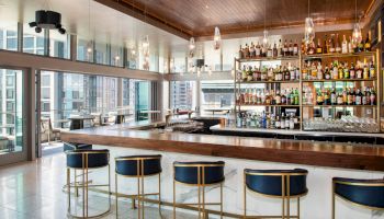 The image shows a modern bar with high stools, a large selection of bottles, and floor-to-ceiling windows offering a city view. The sentence ends here.