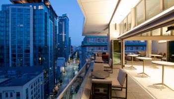 A modern urban balcony with seating overlooks a city street flanked by tall buildings, with an open indoor area visible.
