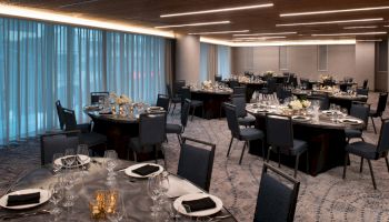 A well-lit, elegant banquet hall with round tables set for dinner, featuring black chairs, neatly arranged tableware, and floral centerpieces.