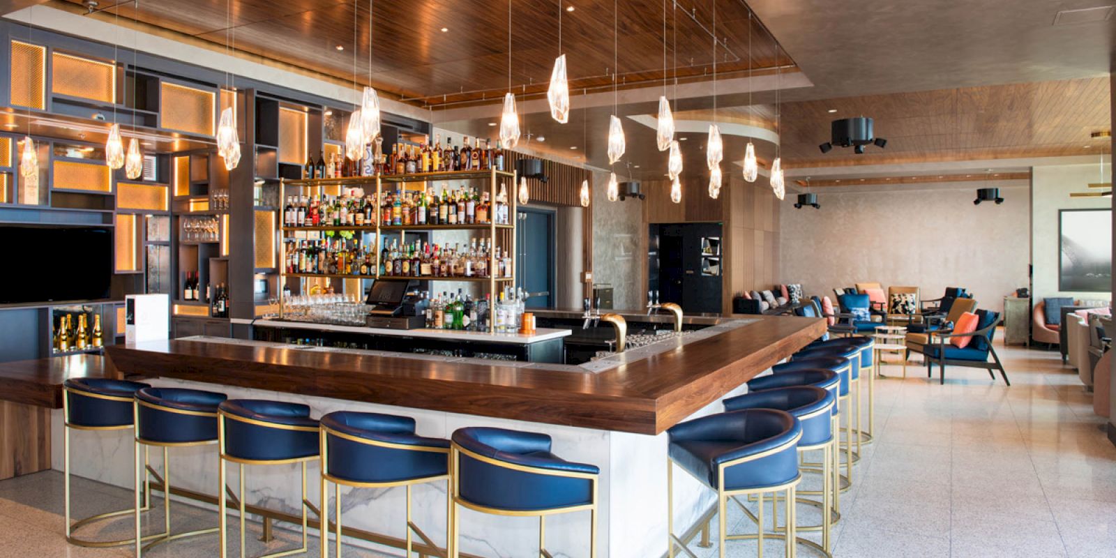 A modern bar with blue stools, a fully stocked shelf of liquor, and hanging lights over a wooden counter. Lounge seating is in the background.
