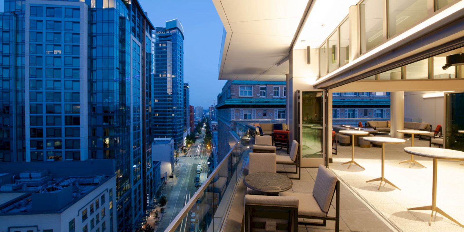 A modern outdoor terrace at dusk with seating areas, overlooking a cityscape featuring high-rise buildings and street below, gently lit.