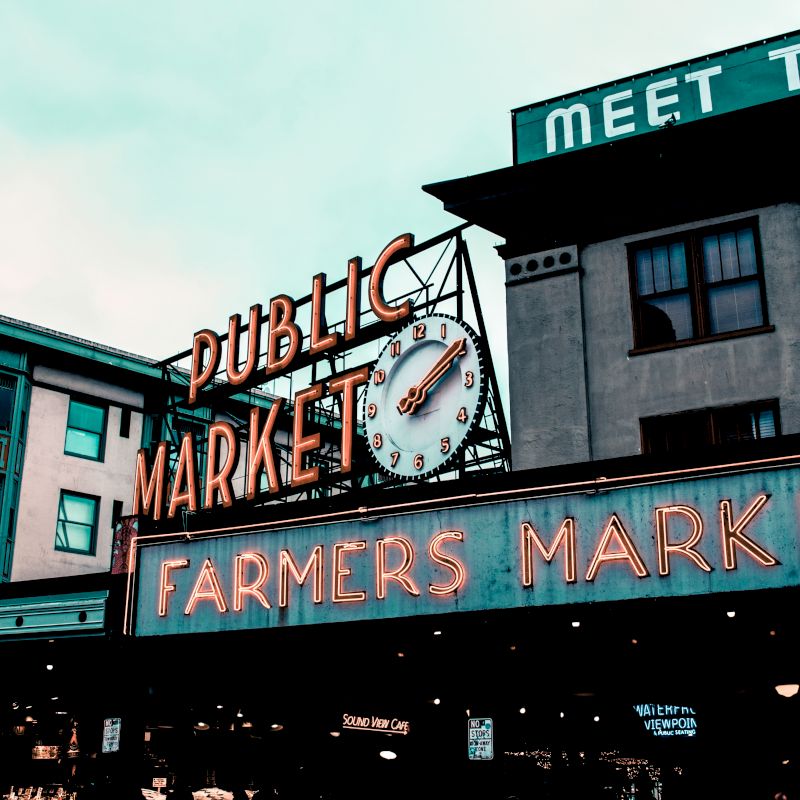 The image shows a public market with a large neon sign reading "Public Market" and "Farmers Market" beneath it, with buildings in the background.