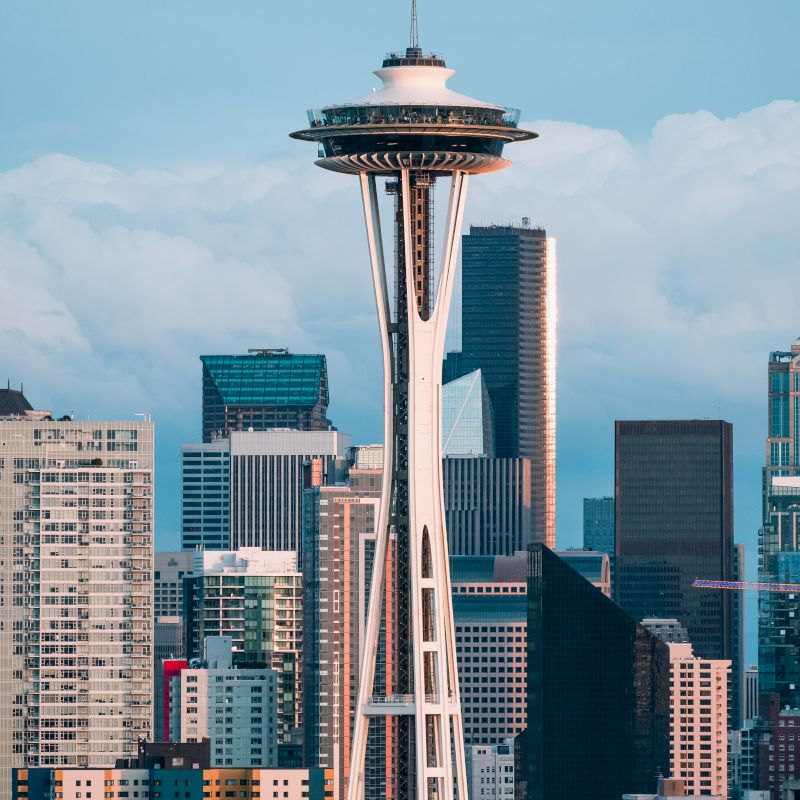 The image shows the Seattle Space Needle with a backdrop of various high-rise buildings in the city skyline on a clear day, creating a vivid urban scene.
