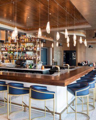 This image shows a modern bar with a U-shaped counter, blue high chairs, and hanging lights, featuring a fully stocked back bar and lounge area.