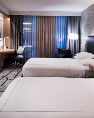 A modern hotel room with two beds, a television on the wall, a desk with a chair, and large windows with curtains.