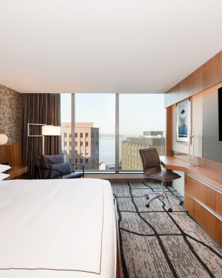 A modern hotel room features a large bed, an armchair, a desk with a chair, and a flat-screen TV on a wooden console, with a city view window.