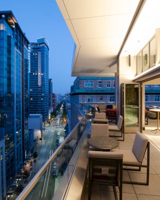 A modern outdoor balcony with seating and tables overlooking a cityscape, featuring tall buildings and a dimly lit blue sky in the evening.