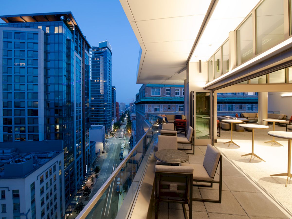 The image shows a modern cityscape with tall buildings, viewed from a high-rise balcony decorated with tables and chairs under warm evening light.