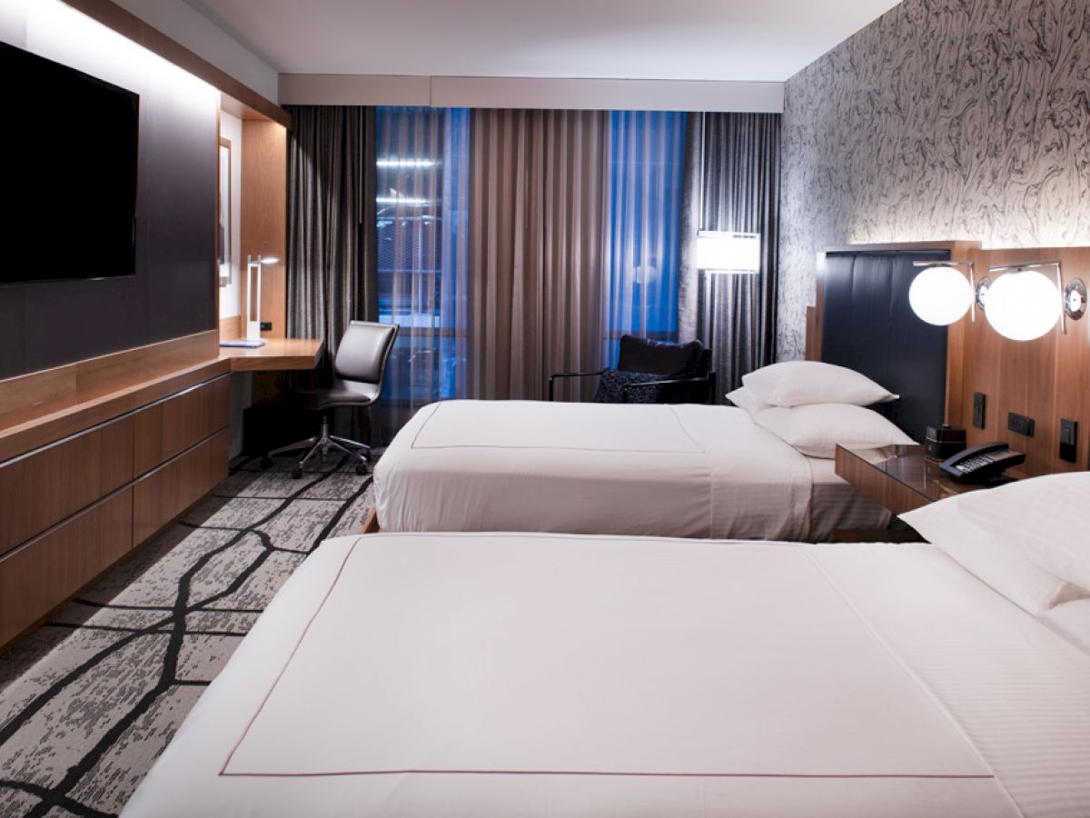 A modern hotel room with two double beds, a wall-mounted TV, a desk with a chair, and large windows with curtains.