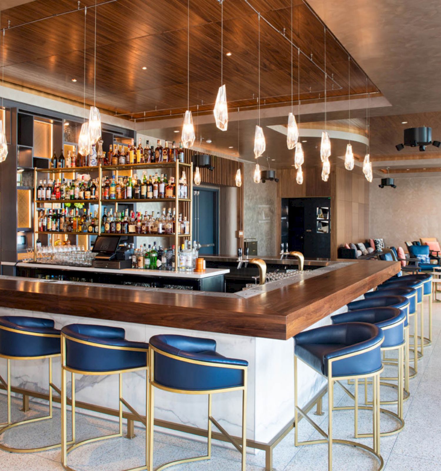 This image shows a modern bar with a well-stocked shelf of liquor bottles, stylish seating, hanging lights, and a cozy seating area in the background.