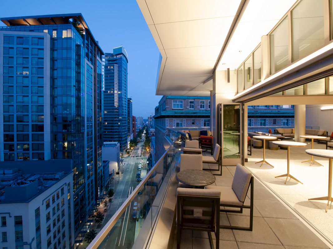 A modern balcony view of a cityscape at dusk, with tall buildings and an outdoor seating area with tables and chairs.