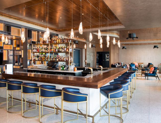 This image shows a modern bar area with a counter, multiple bar stools, hanging lights, and a fully stocked bar in the background, ending the sentence.