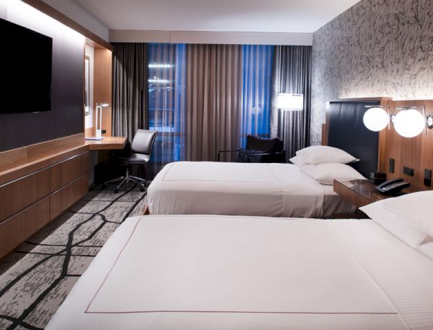 A modern hotel room with two neatly made beds, a TV mounted on the wall, a desk and chair, and stylish lighting and decor on the walls.