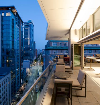 A modern cityscape view from a balcony at dusk, with outdoor seating and tables flanking the glass railing along a high-rise building.