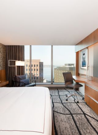 A modern hotel room with a large bed, desk, chair, wall-mounted TV, and large window showing a view of buildings and water.