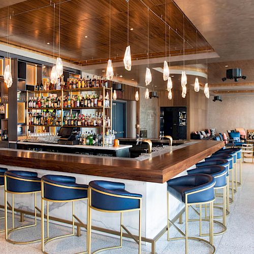The image shows a stylish bar area with a wooden counter, blue cushioned bar stools, liquor bottles on shelves, and contemporary pendant lights.