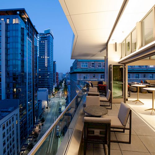 A modern balcony with seating and tables, overlooking a city street with tall buildings, illuminated at dusk.