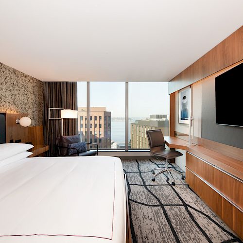 A modern hotel room with a large bed, a TV, desk, chair, and a view of buildings and water through a large window ending the sentence.