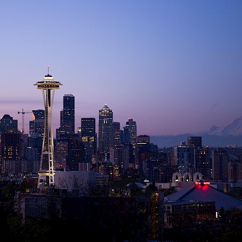 A cityscape during twilight featuring the Space Needle, high-rise buildings, and Mount Rainier in the background with a clear sky.