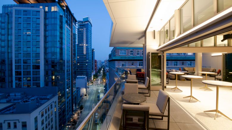 The image shows a modern cityscape with tall buildings and a balcony at dusk, featuring outdoor seating and tables on the right.