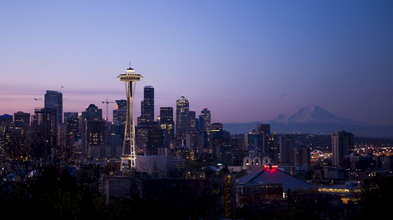 This image shows the skyline of Seattle at dusk, including the Space Needle and Mount Rainier in the background, with city lights illuminating the scene.