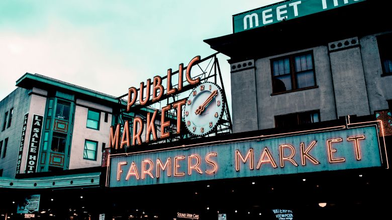 The image shows a public market with a neon sign that reads "Farmers Market" and a clock. The surrounding buildings include a hotel.