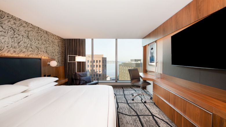 A modern hotel room with a large bed, wall-mounted TV, desk, chair, and a window offering a city view. The room has a patterned wall and carpet.
