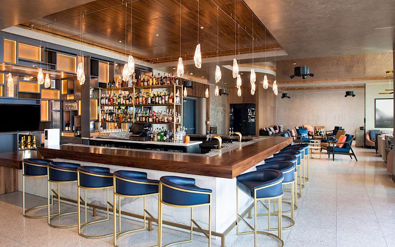 This image shows a modern bar with a well-stocked shelf, blue and gold bar stools, hanging lights, and a seating area in the background.