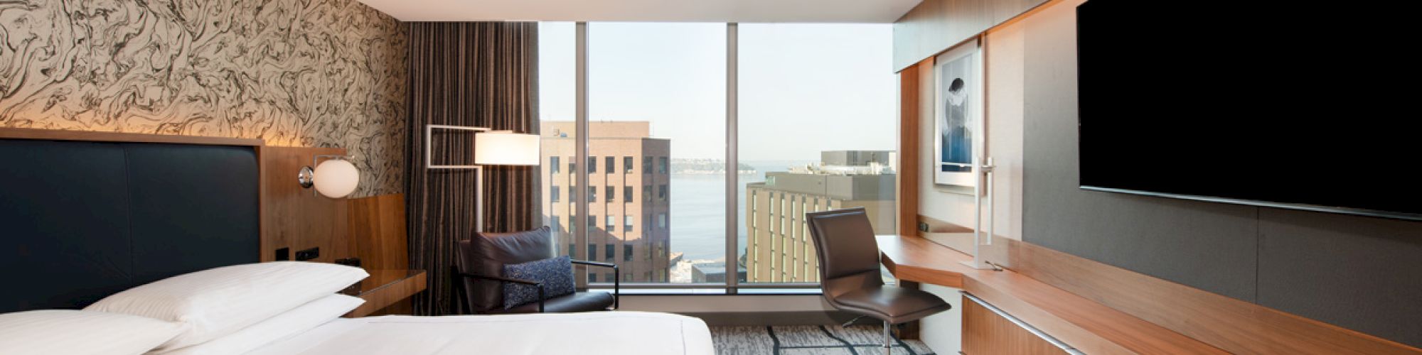 A modern hotel room with a large bed, a flat-screen TV, a desk, and a view of buildings through a large window.