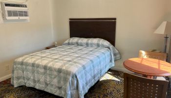 A simple room with a bed, table, chairs, lamp, and wall-mounted air conditioner on a patterned carpet.