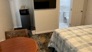 A hotel room with a bed, round table, chair, wall-mounted TV, mini fridge, closet, and a bathroom visible through an open door.